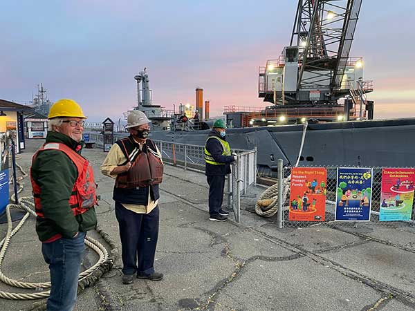 three guys in life vests and hard hats in front of Pampanito and crane barge in background.